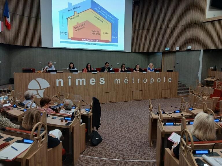 Salle du conseil de Nîmes Métropole lors d’une réunion publique, avec plusieurs intervenants assis derrière une grande tribune en bois sous un écran de présentation projeté. Les participants sont installés dans un amphithéâtre moderne aux tons bois et gris.