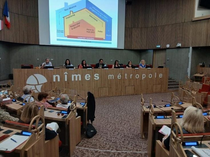 Salle du conseil de Nîmes Métropole lors d’une réunion publique, avec plusieurs intervenants assis derrière une grande tribune en bois sous un écran de présentation projeté. Les participants sont installés dans un amphithéâtre moderne aux tons bois et gris.