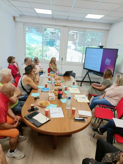 Photographie d'une rencontre organisée autour d'une table ovale, les personnes présentes regardent le support de formation et écoutent les commentaires de la formatrice